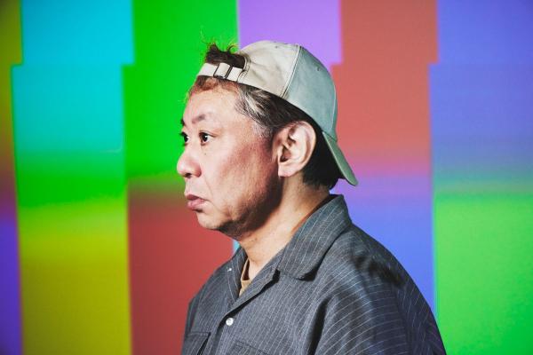 カラーバーのようなカラフルな背景の前にいる鈴木おさむの横顔の写真